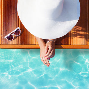 A woman wearing sunglasses and a hat enjoys her time on a wooden deck next to a refreshing swimming pool.